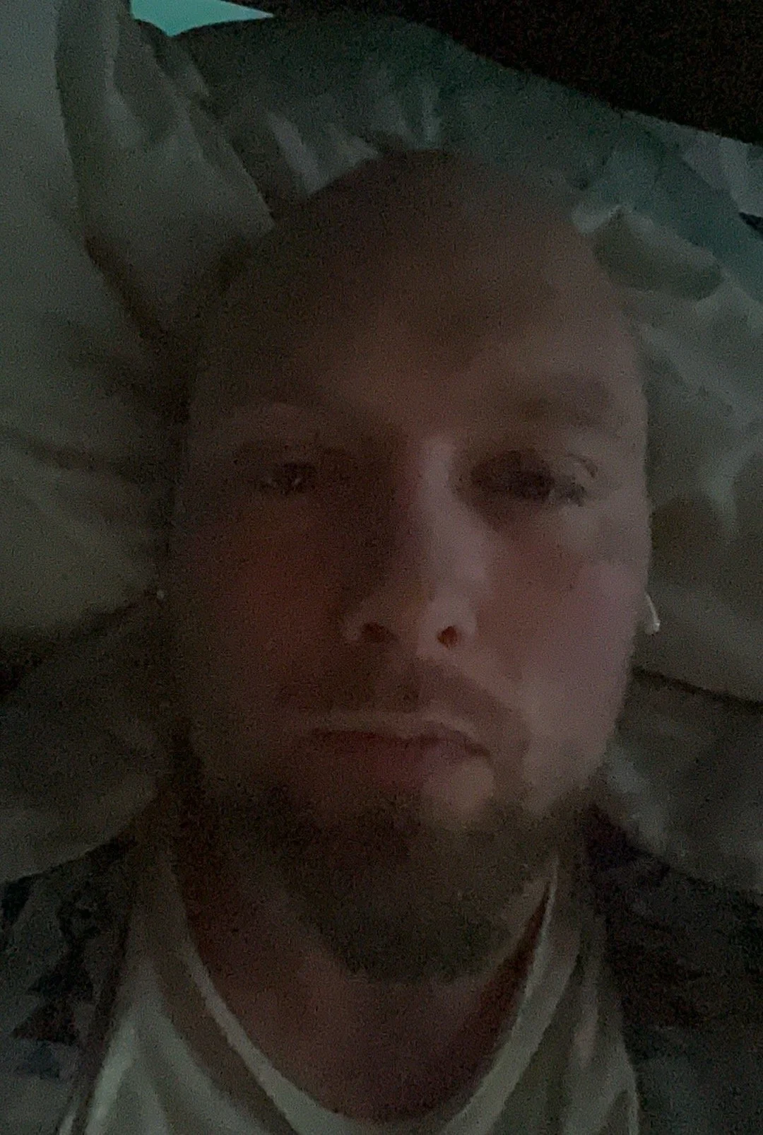 A person with a shaved head and earrings lying down on a pillow in a dimly lit room.
