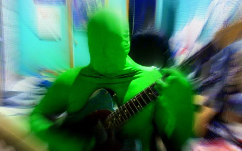 A person wearing a green full-body suit playing an acoustic guitar in a cluttered room.