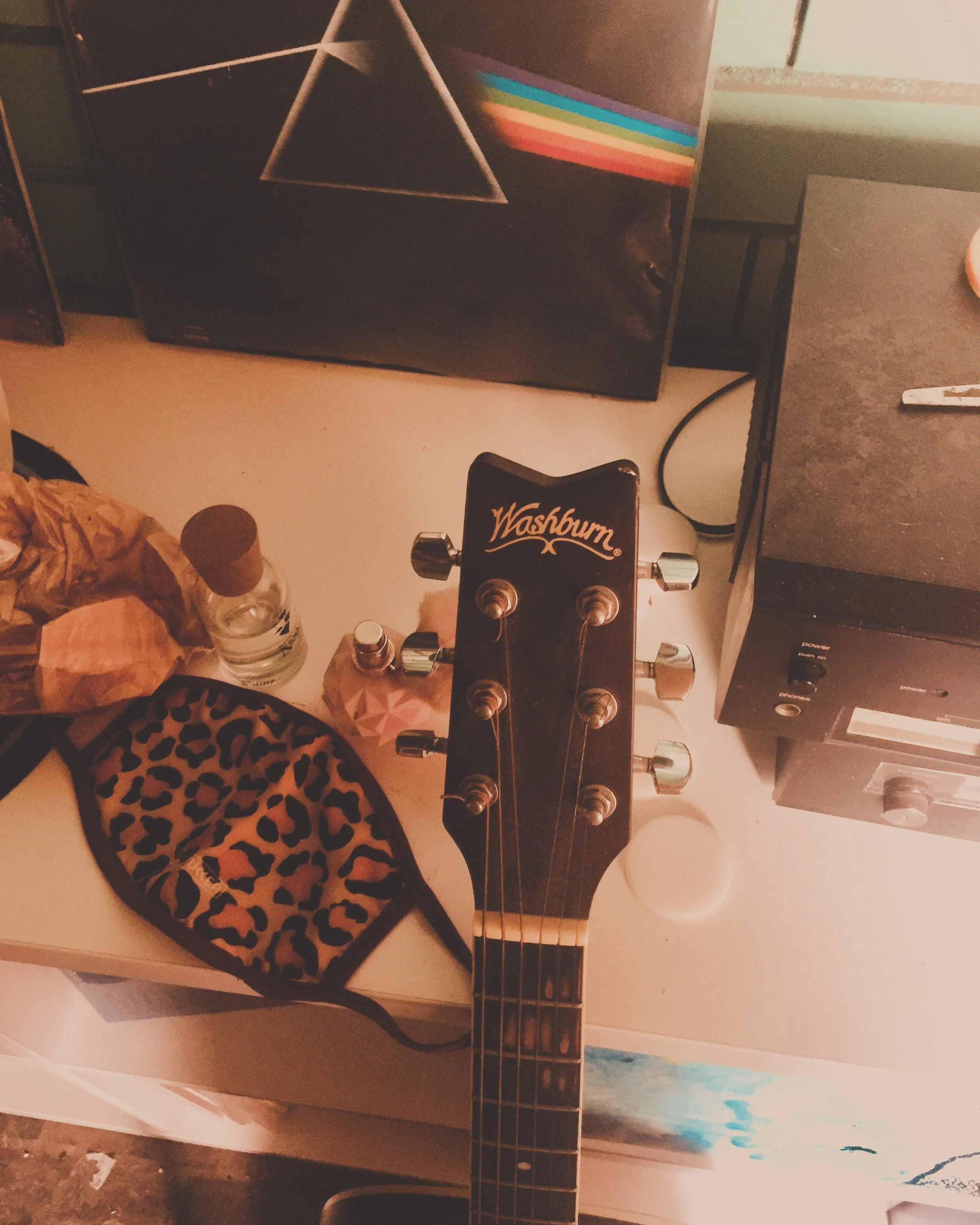Close-up of a Washburn guitar headstock with tuning pegs, resting on a white surface. Surrounding objects include a leopard print cloth, a small bottle, a pink geometric object, a brown paper bag, and electronic equipment, with a framed object featur