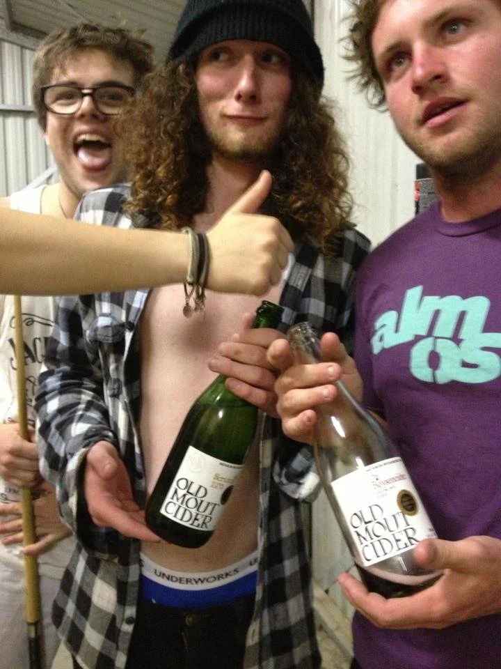 Three young men at a party, two holding bottles of cider labeled 'Old Mount Cider.' The man in the middle is wearing a beanie and plaid shirt, giving a thumbs-up, while the other two are smiling and holding the bottles.