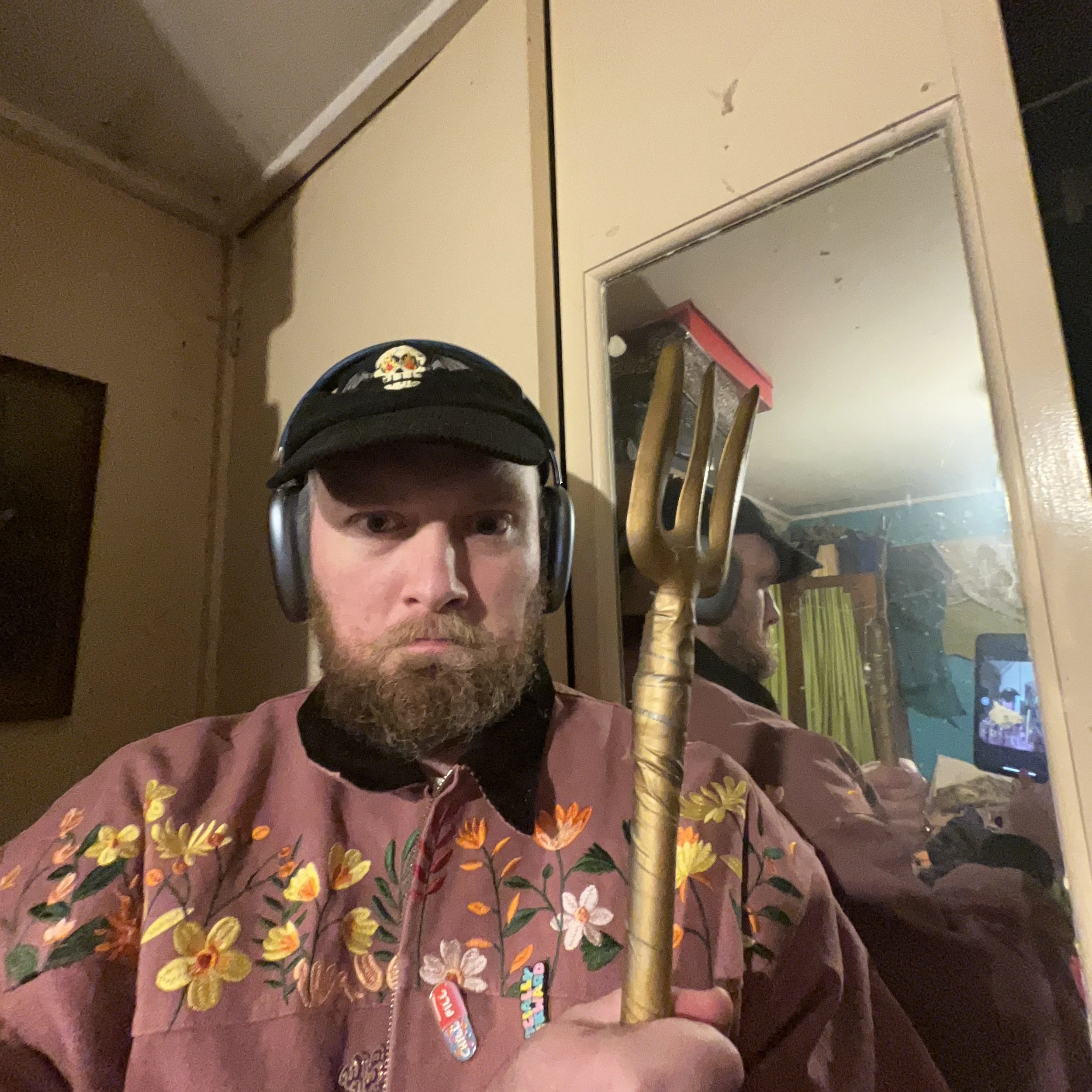 A man with a beard wearing a black cap with a skull patch, headphones, and a pink floral jacket, holding a small gold-colored rake in front of a mirror, which reflects his profile and a room with a computer monitor.