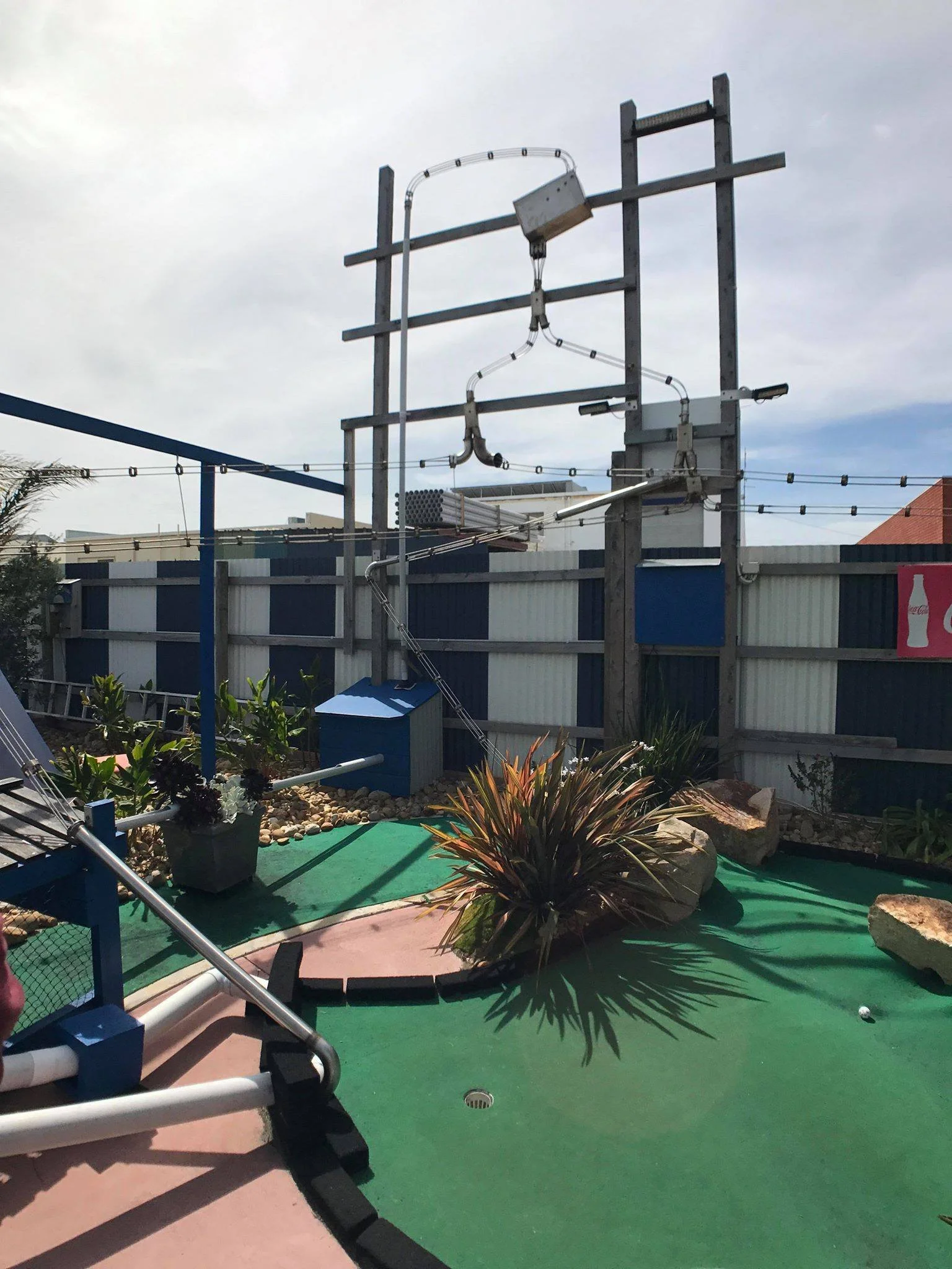 Miniature golf course with a windmill obstacle, plants, rocks, and a colorful surface under a partly cloudy sky.