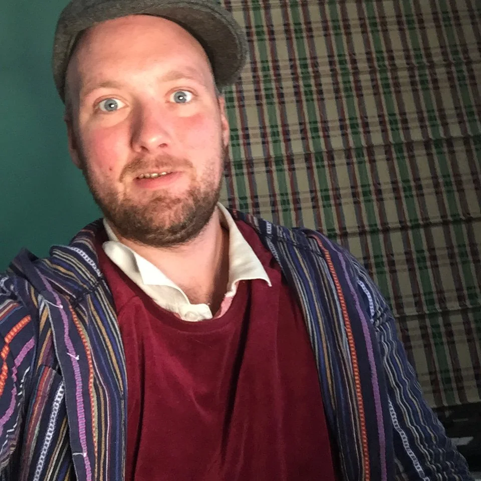 A man with a beard and mustache wearing a gray hat, a white shirt, a red velvet shirt, and a multicolored striped jacket, taking a selfie in front of a plaid pattern backdrop.