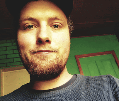 A young man with a beard and mustache smiling indoors with green walls and a framed picture in the background.