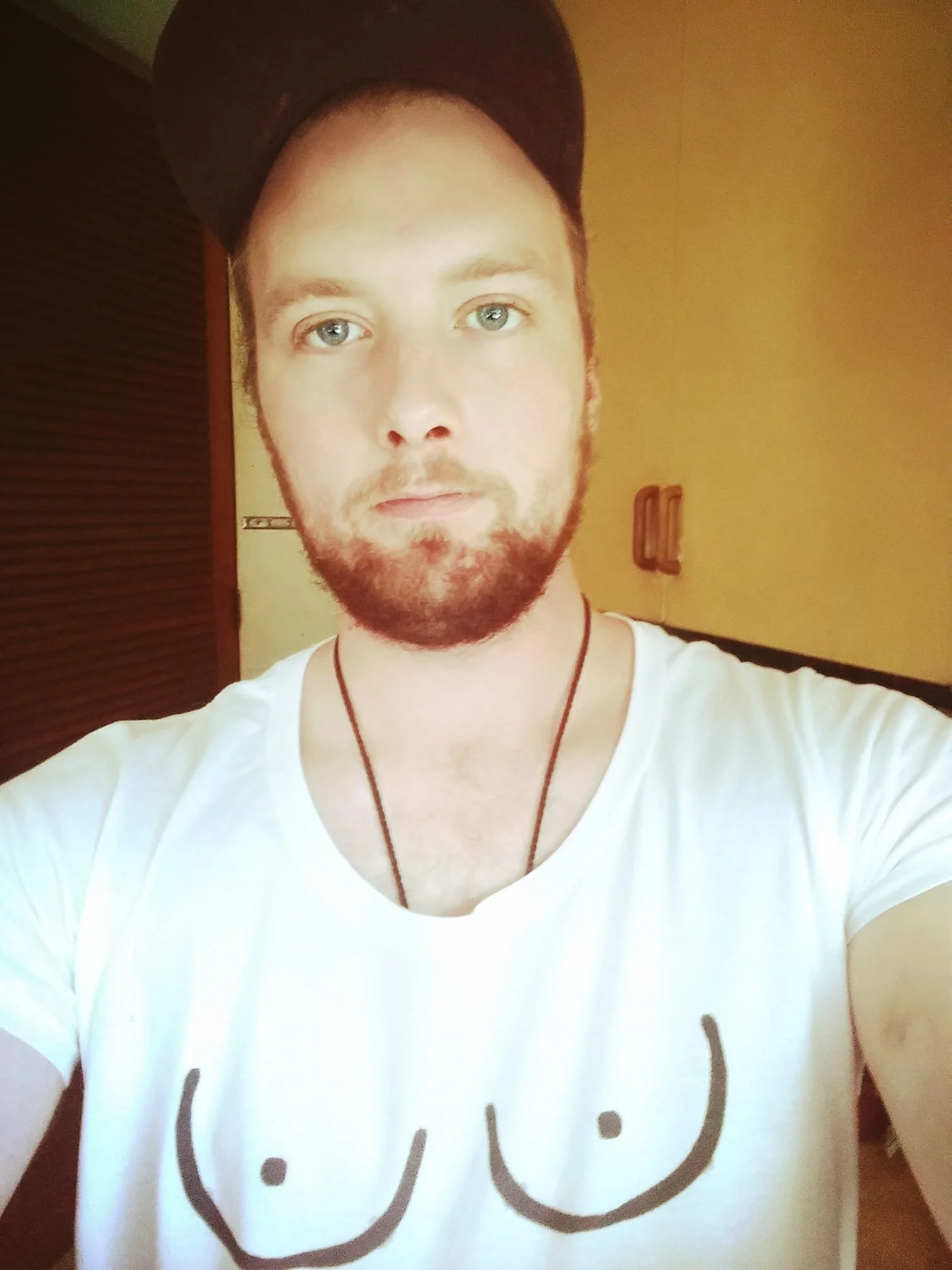 A young man taking a selfie indoors, wearing a white t-shirt with a smiley face print and a black cap. He has light skin, blue eyes, a beard, and a necklace.