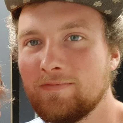 Close-up of a young man with light skin, blue eyes, reddish-brown beard, and mustache wearing a patterned hat.