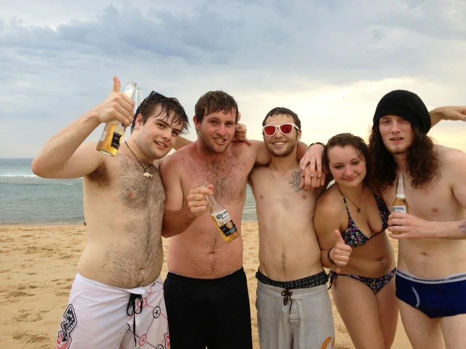 Group of five young adults at the beach, shirtless, holding beers, and smiling.