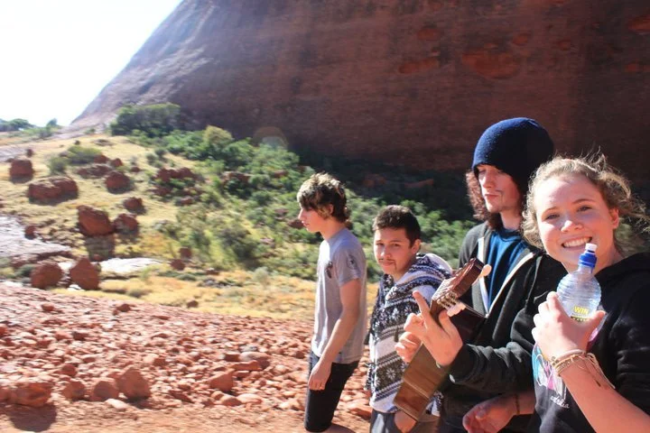 Four kids walking outdoors in a desert landscape with red rocks and a large rock formation in the background. One girl is smiling and holding a water bottle, the second girl is holding a guitar, and the two boys are walking behind them.