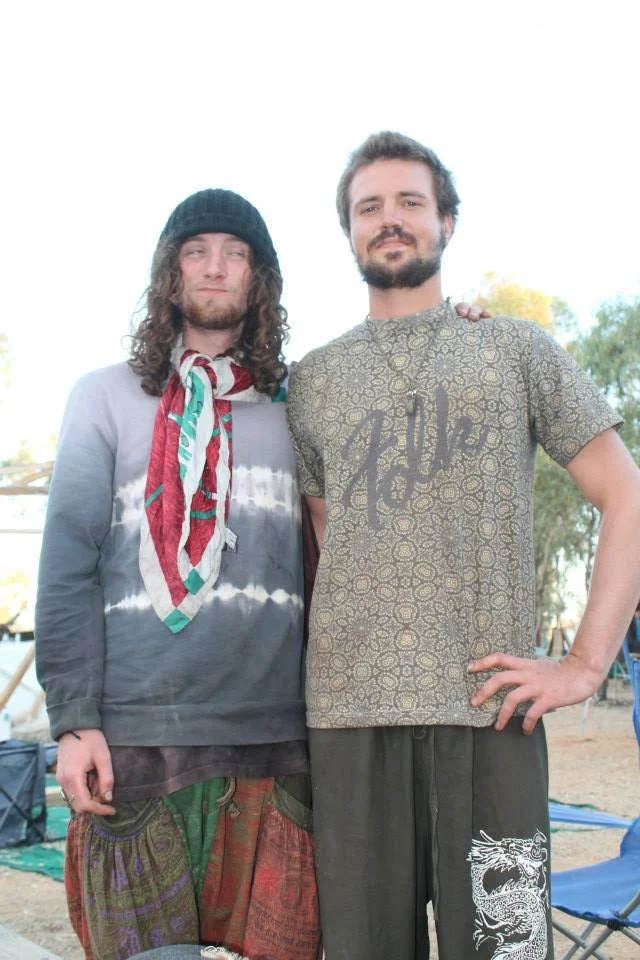 Two men standing outdoors, one wearing a beanie and a tie-dye long-sleeve shirt, the other in a patterned t-shirt and shorts, with trees and outdoor furniture in the background.