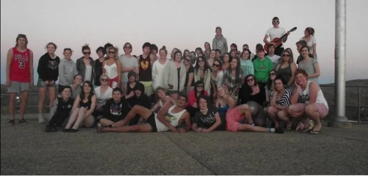 Group of young people posing outdoors on a paved area, with a cloudy sky in the background. Some are sitting, others standing, with one person playing an electric guitar.