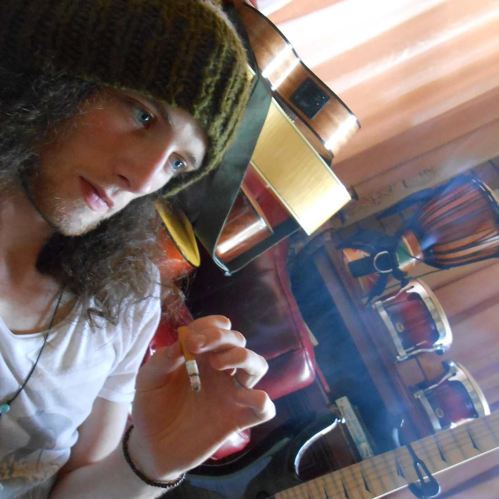 A man with long curly hair, wearing a beanie, sitting in a music or entertainment venue, holding a cigarette.