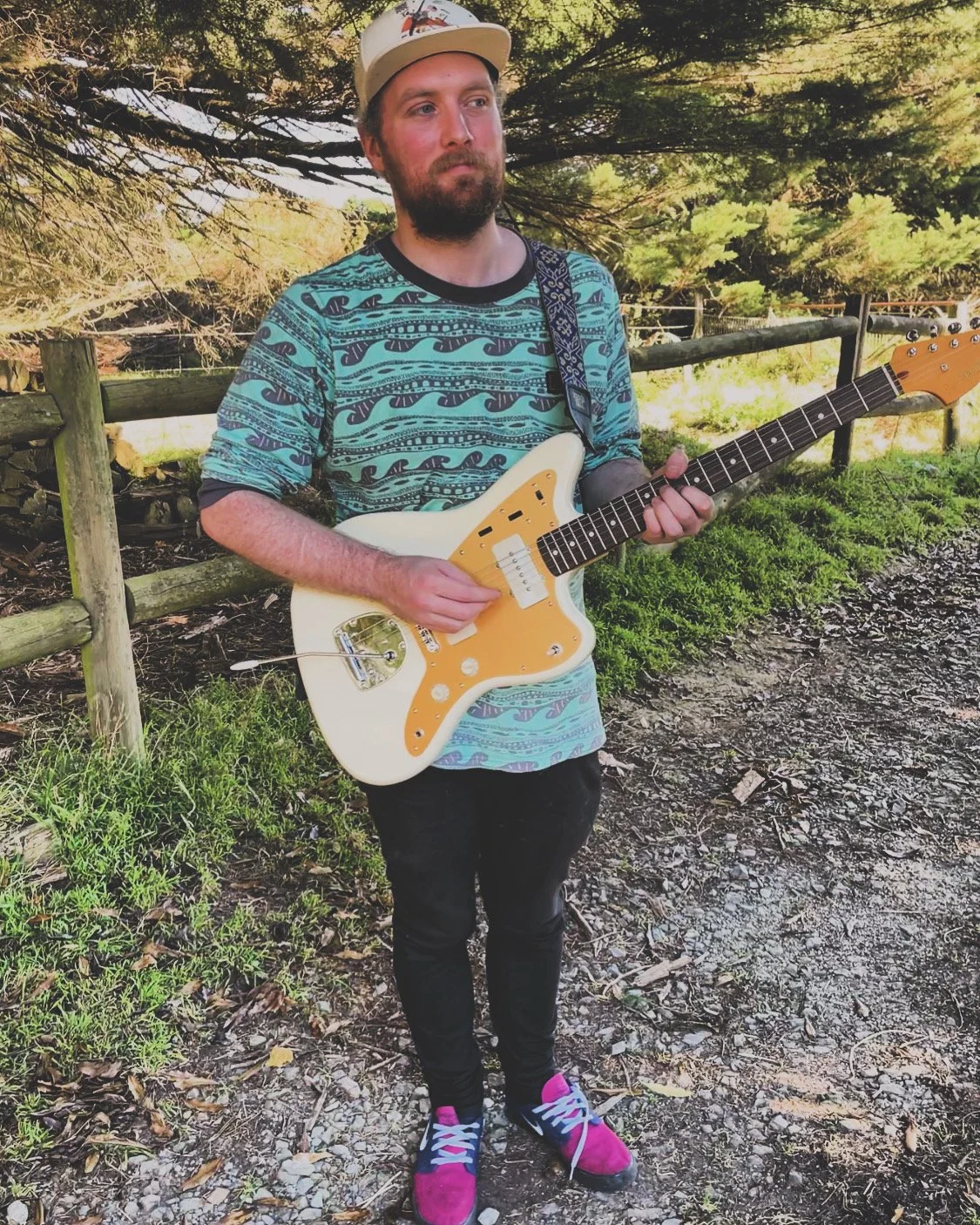 A man with a beard, wearing a baseball cap and a blue patterned t-shirt, is standing outdoors holding a white electric guitar with an orange pickguard.