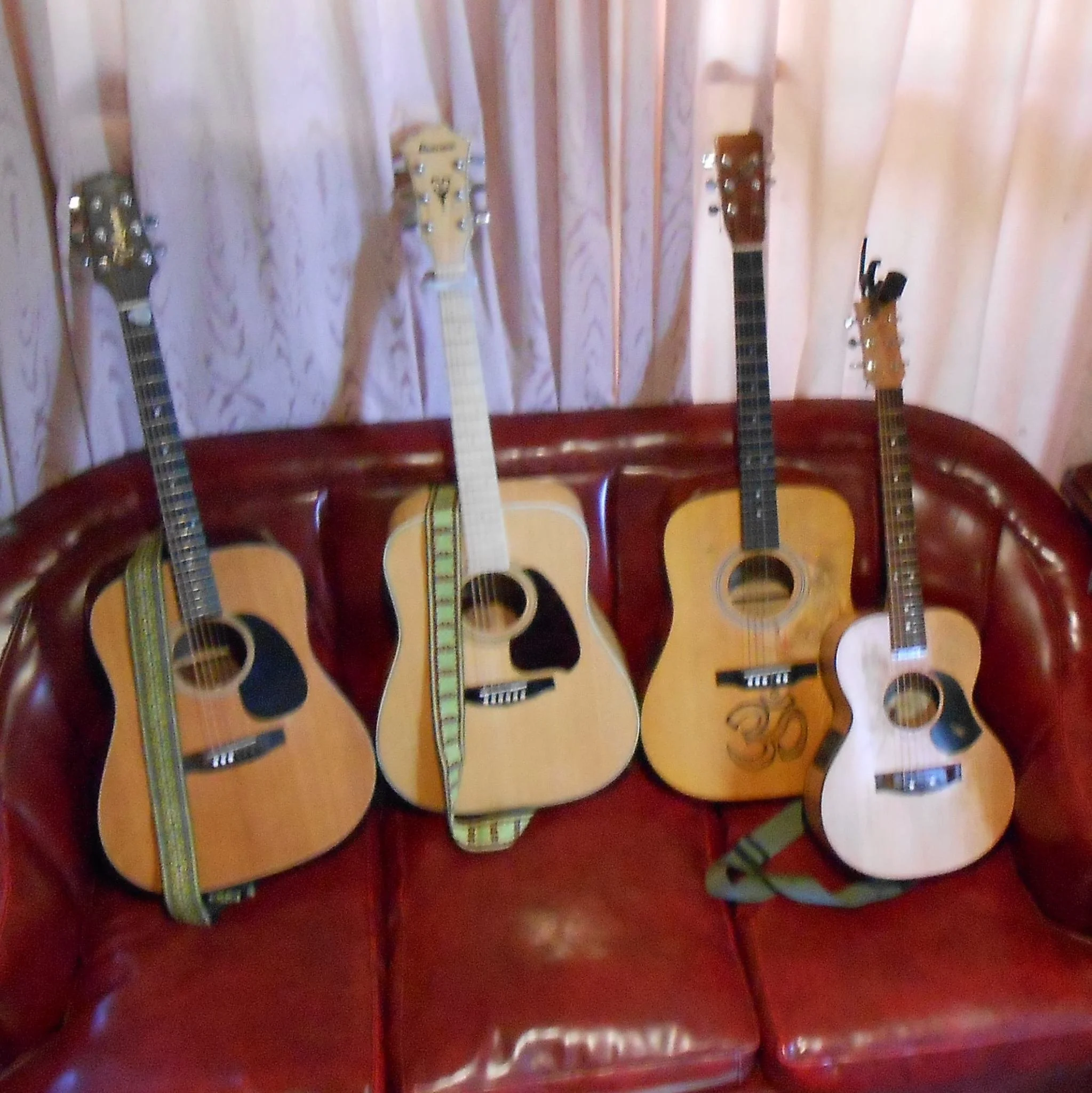 Four acoustic guitars resting against a red leather couch in front of pink curtains.