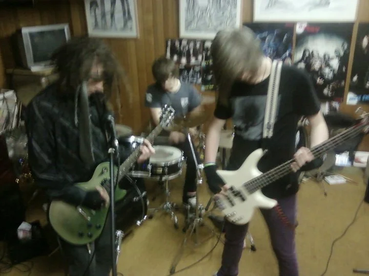 Three young musicians playing guitars and drums in a room with wood-paneled walls and posters.