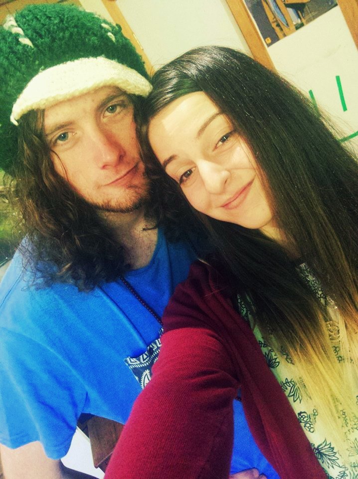 A young man with long curly hair wearing a green and white knit hat and a blue shirt, taking a selfie with a young woman with long straight black hair, both smiling in an indoor setting.