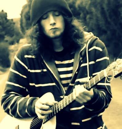 Person playing an acoustic guitar outdoors, wearing a beanie, striped shirt, and hoodie.