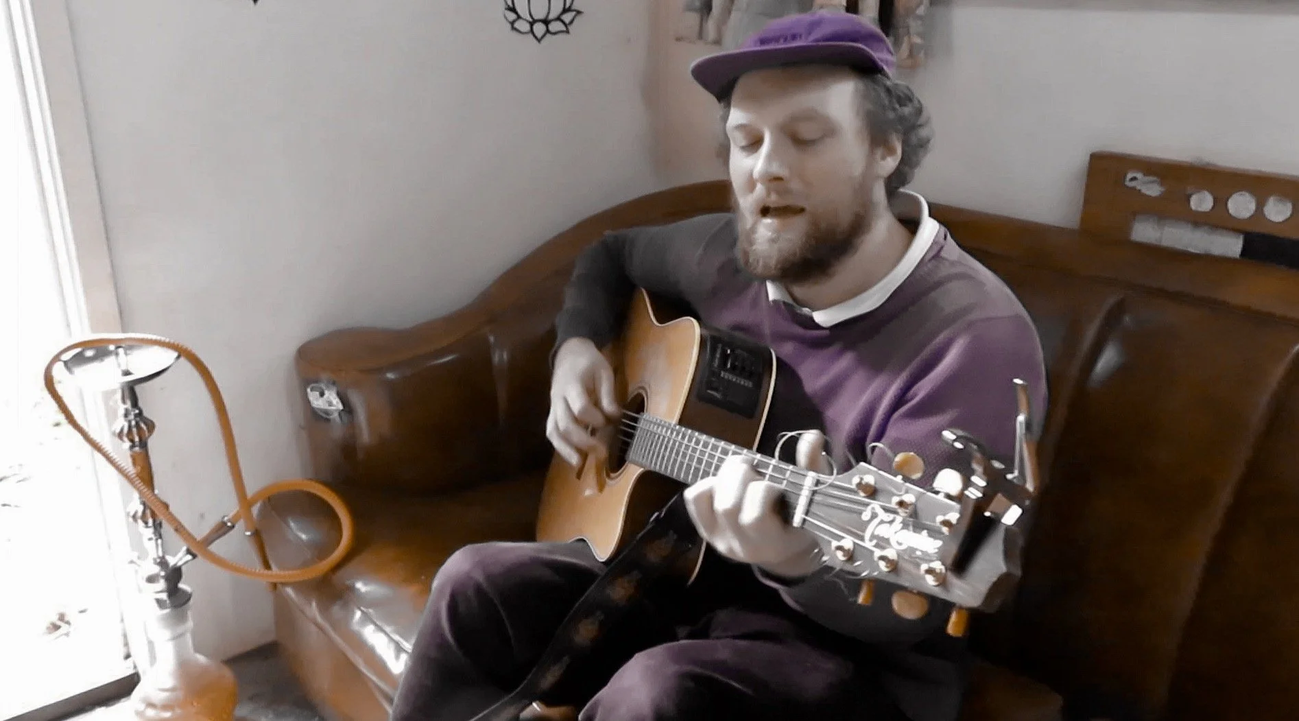 A man with a beard wearing a purple cap and a purple and white shirt playing an acoustic guitar while sitting on a wooden couch in a room with a lamp and wall decorations.