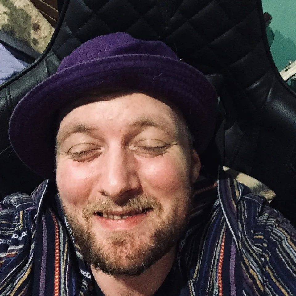 A man with a beard and mustache smiling with his eyes closed, wearing a dark purple hat and a multi-colored striped shirt, sitting in a black chair.