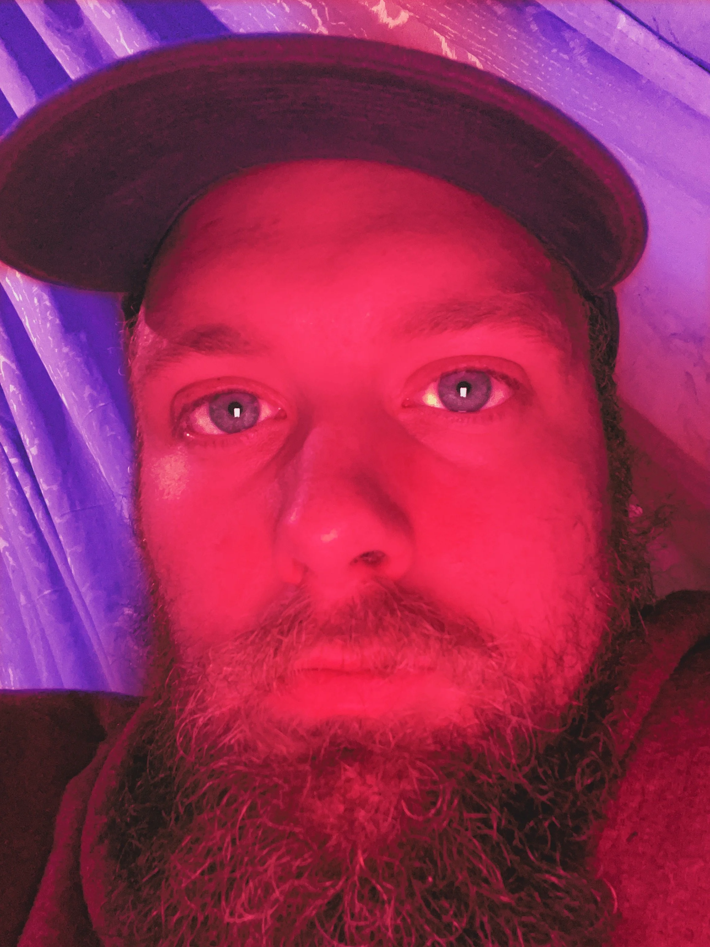 Close-up of a man with a beard and mustache, wearing a cap. His face is lit with a red/pink light, and he has blue eyes. The background includes a curtain with a pattern.