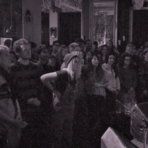 Crowd of people gathered indoors, likely at a concert or event, with a woman in the foreground looking towards the stage.