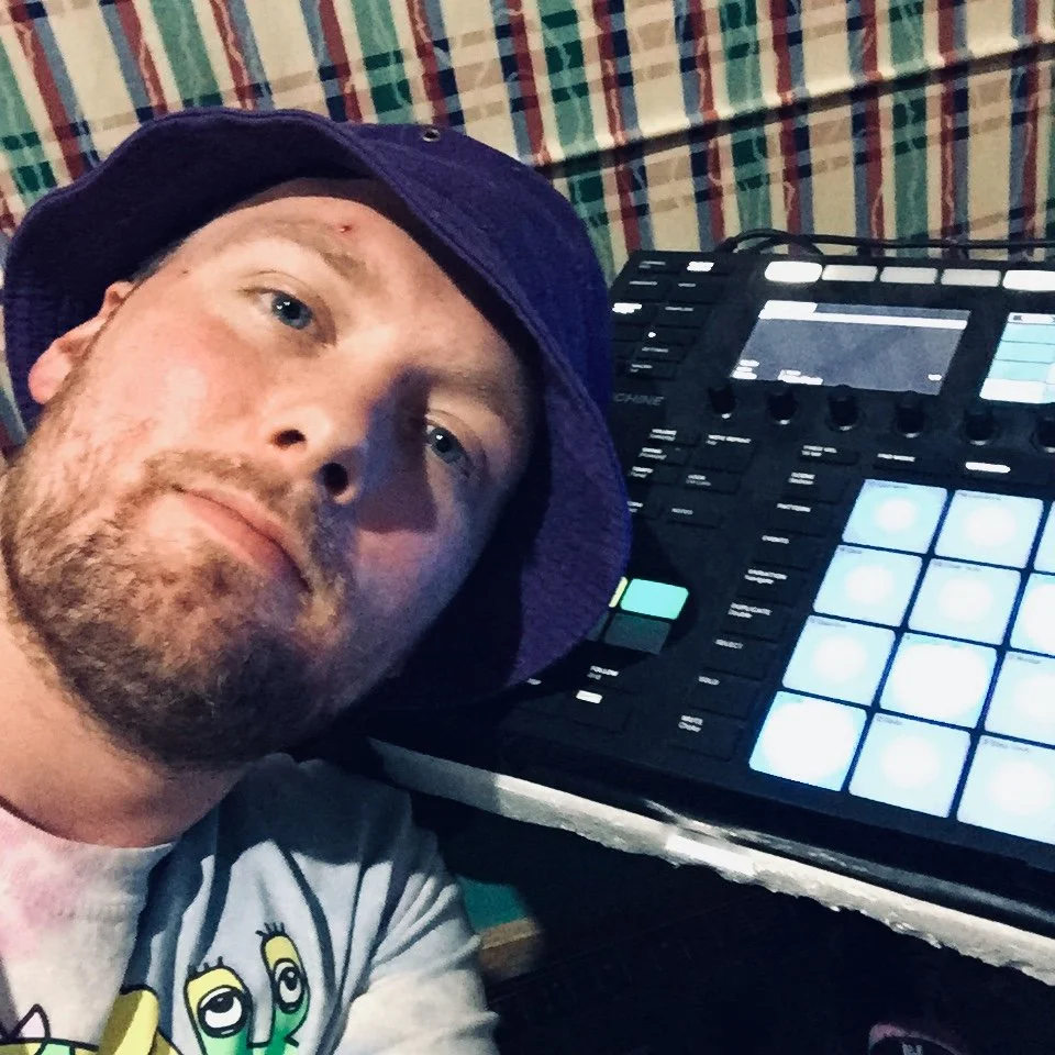 A man wearing a blue hat and a light-colored shirt with cartoon eyes prints, taking a selfie with a music production controller with illuminated pads in the background.