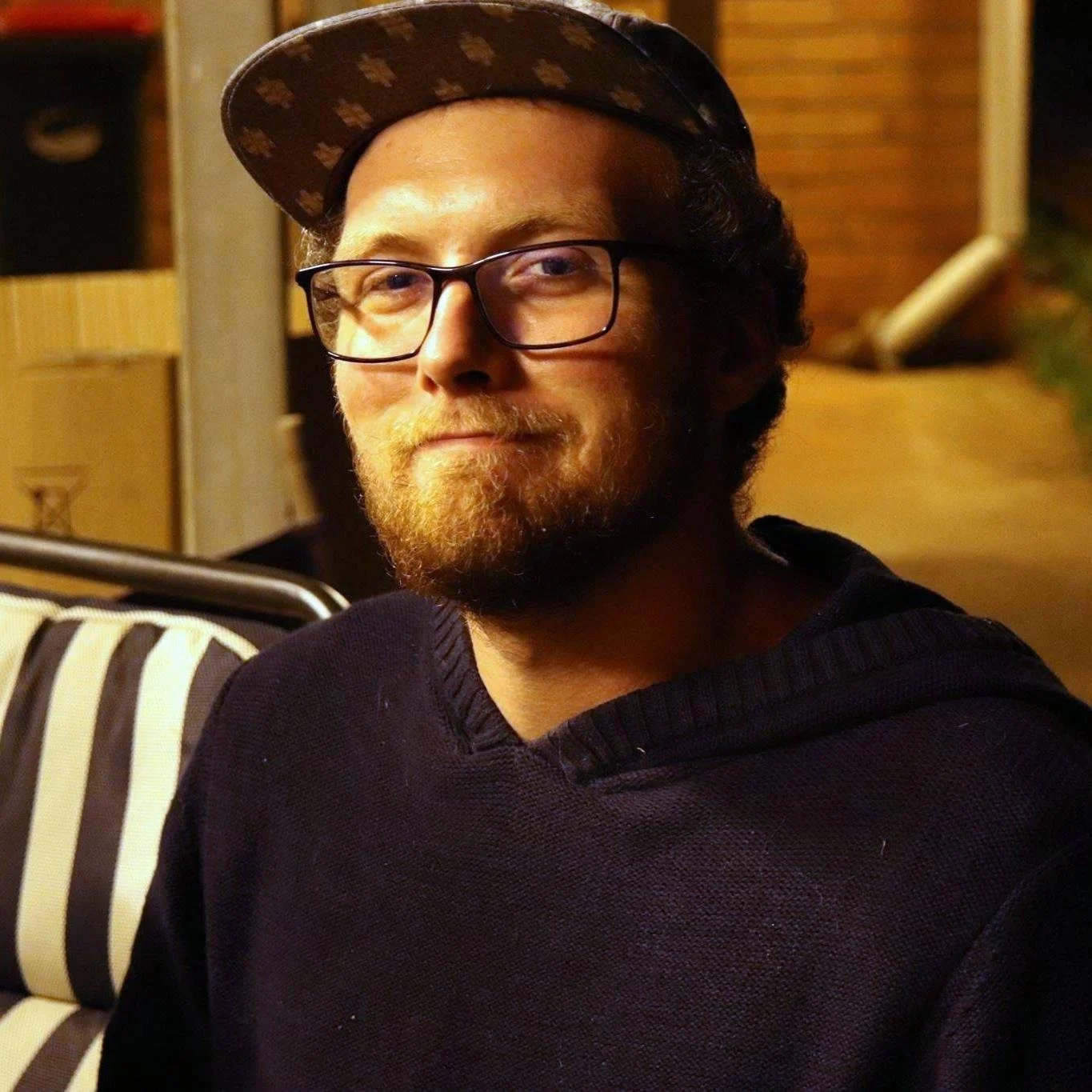 A man with glasses, a beard, and a mustache wearing a dark hoodie and a patterned baseball cap, sitting in a room with wooden and cardboard boxes in the background.