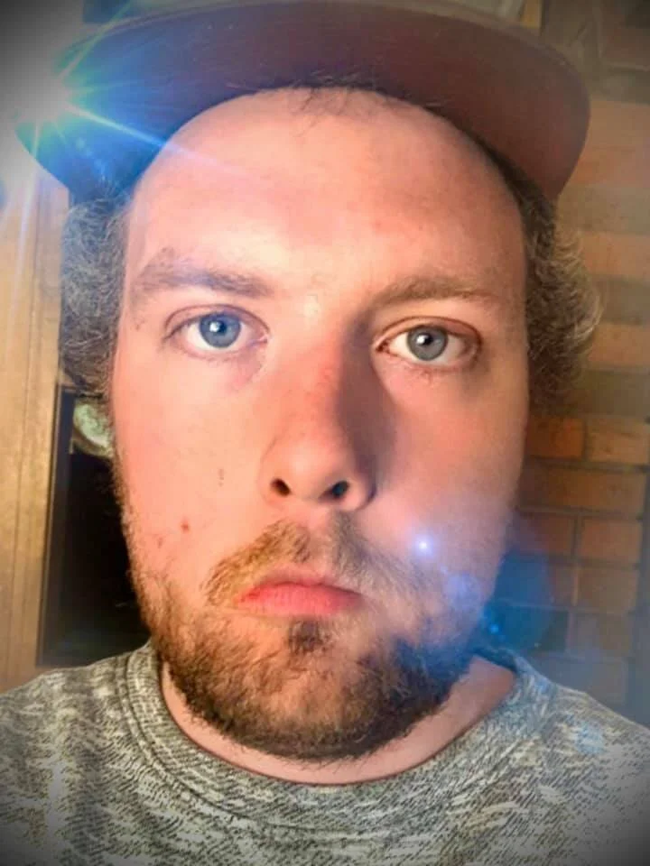 A young man with light skin, blue eyes, and brown, curly hair, wearing a gray hat and a patterned gray shirt, looks directly at the camera with a neutral expression.
