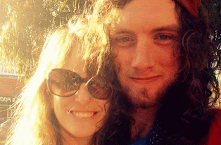 A smiling woman with long hair and sunglasses taking a selfie with a man with long curly hair. They are outdoors in sunlight.