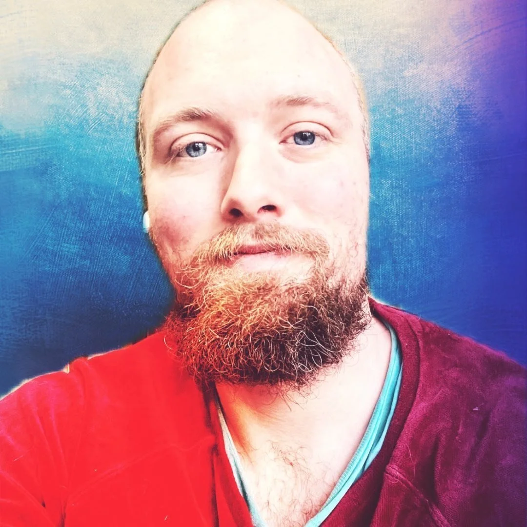 A close-up selfie of a man with a beard and mustache, wearing a red shirt, standing against a colorful background with shades of blue, purple, and pink.