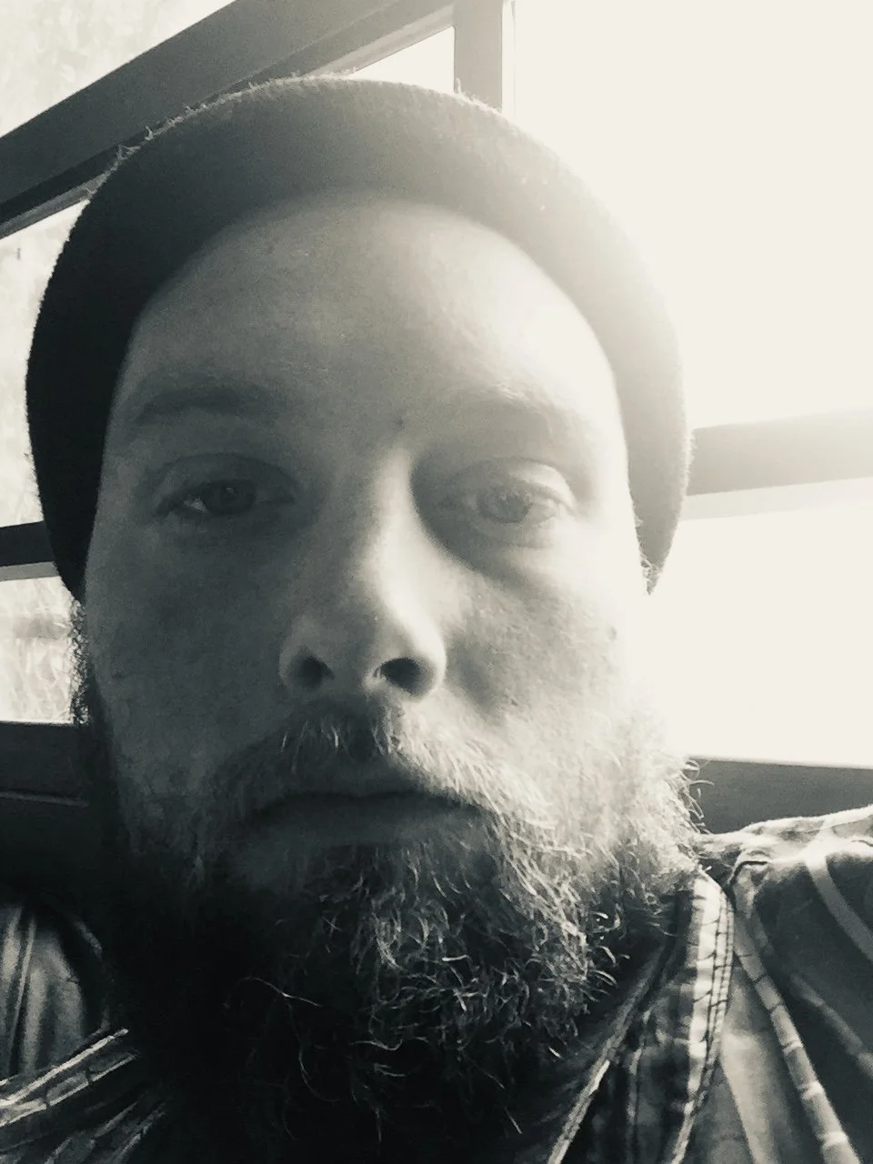 Close-up black-and-white photo of a man with a beard and mustache, wearing a hat, sitting near a window with sunlight streaming in.