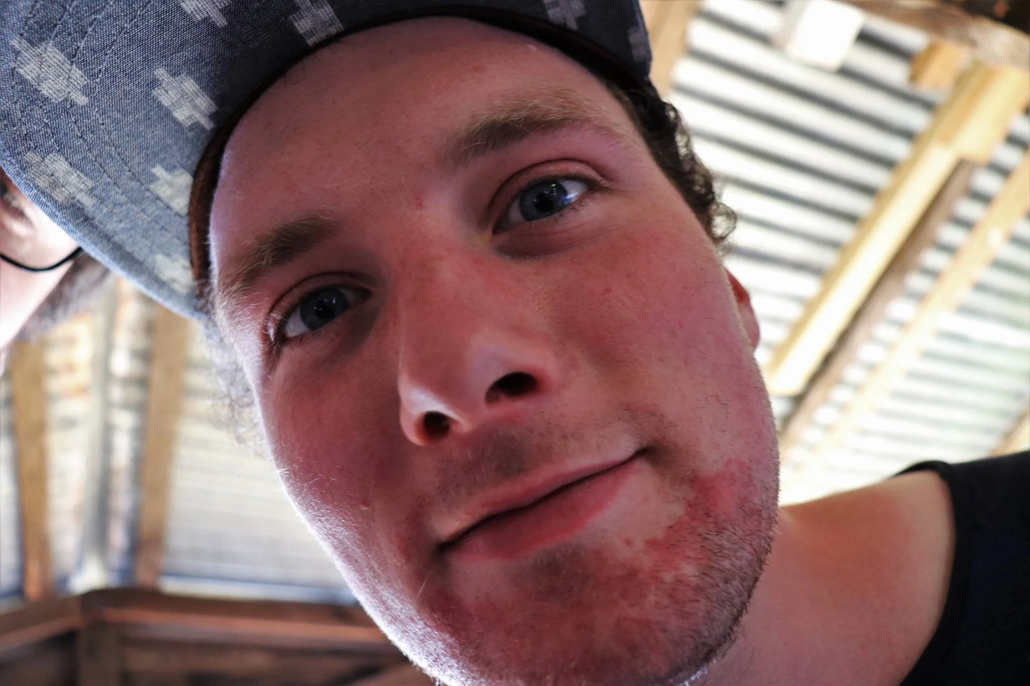 Close-up of a young man's face with light skin, blue eyes, dark hair, and wearing a baseball cap, with a background of a barn or shed ceiling with wooden beams.