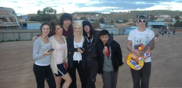 Group of six teenagers standing on a dirt field with buildings and a hill in the background; one is holding a guitar, others are holding phones, and they are dressed casually.