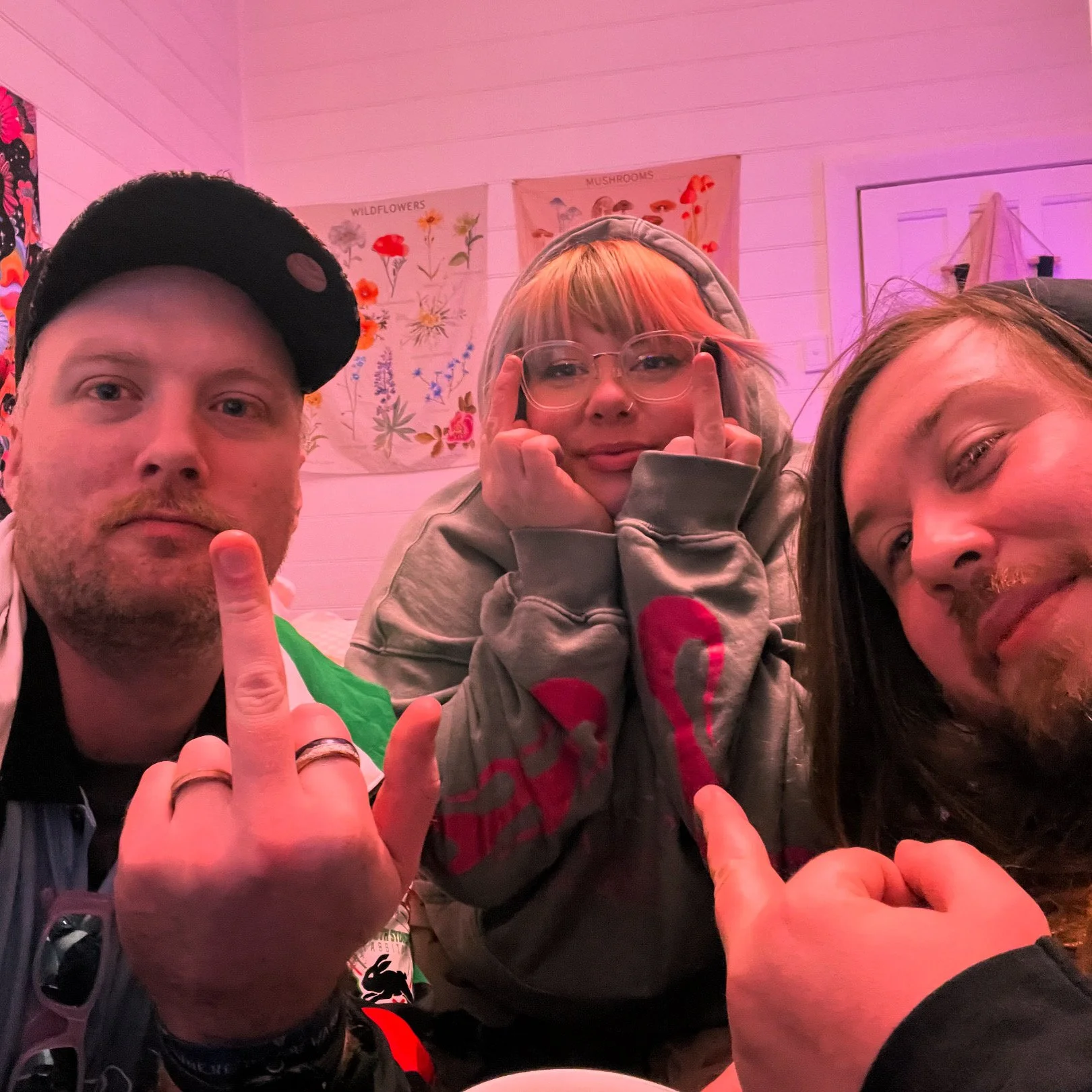 Three people taking a selfie in a room with pink lighting, two men and one woman. The man on the left is wearing a black cap, has a beard, and is giving the middle finger. The woman in the middle has blonde hair, glasses, and is also giving the middl