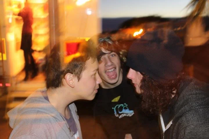 Three young adults interacting outdoors during sunset; two are facing each other closely, and one is behind them with a wide open mouth, expressing excitement.