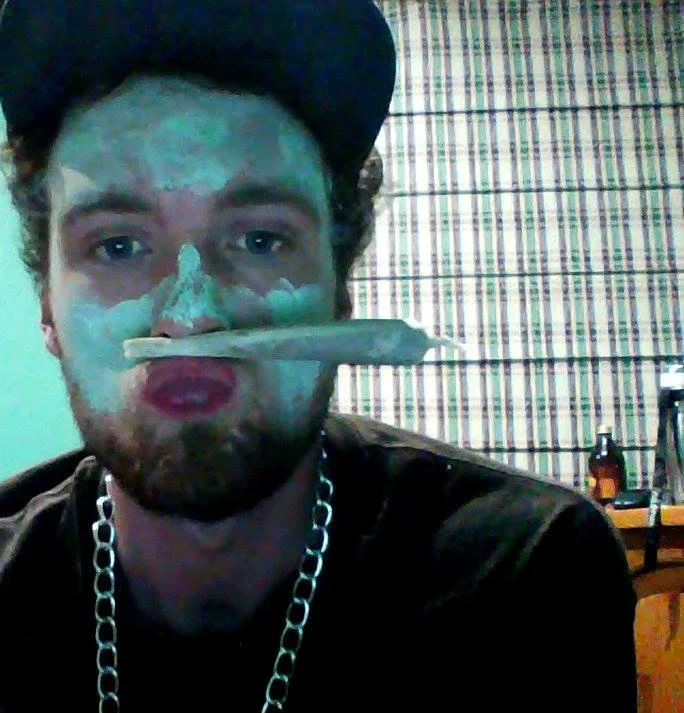 A man with a beard and wearing a black hat makes a humorous face with his lips pursed while holding a rolled joint in his mouth, with lighter green face paint, sitting in a room with a wooden table and a striped curtain.