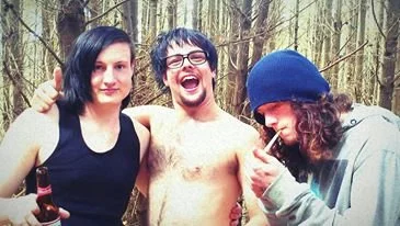 Three young adults outdoors in a forest. The person on the left is a woman with black hair in a black tank top. The person in the middle is a shirtless man with glasses, smiling and laughing. The person on the right is a woman with curly hair, wearin