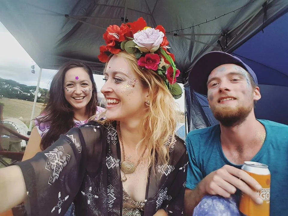 Three people at an outdoor event under a tent, two women and one man, smiling and posing for a selfie. The woman in the center wears a flower crown and has glitter and face paint on her face. The woman on the left has face paint and is smiling. The m