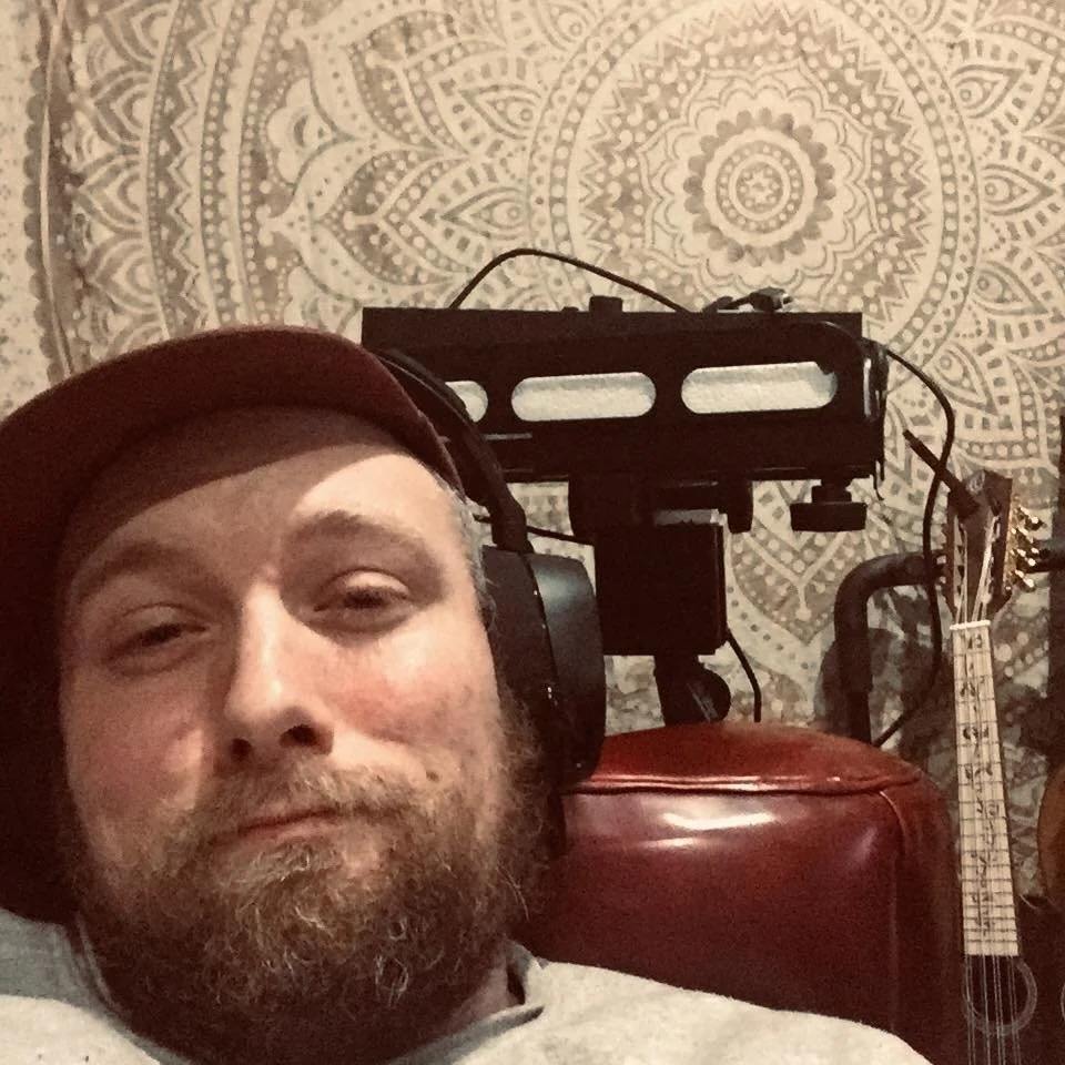 Close-up of a man with a beard, wearing a hat and headphones, taking a selfie indoors. In the background, there is an acoustic guitar, a red leather suitcase, and a mandolin or small string instrument, with a decorative mandala-patterned wall hanging