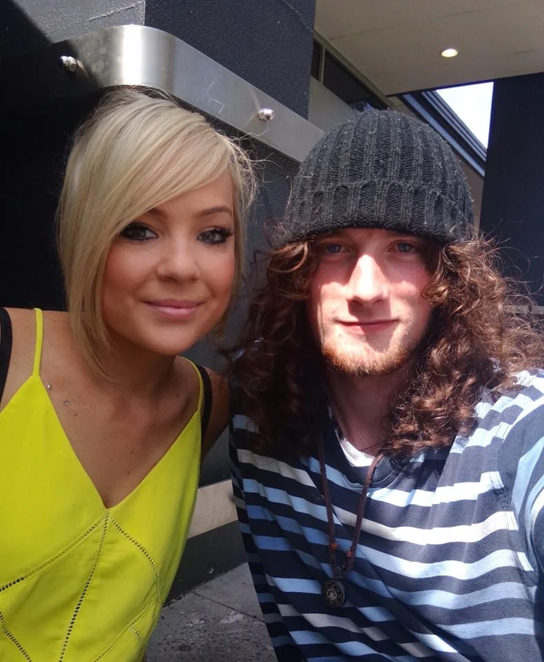 A young woman with blonde hair smiling and a young man with long curly hair wearing a beanie, both taking a selfie outdoors near a building.