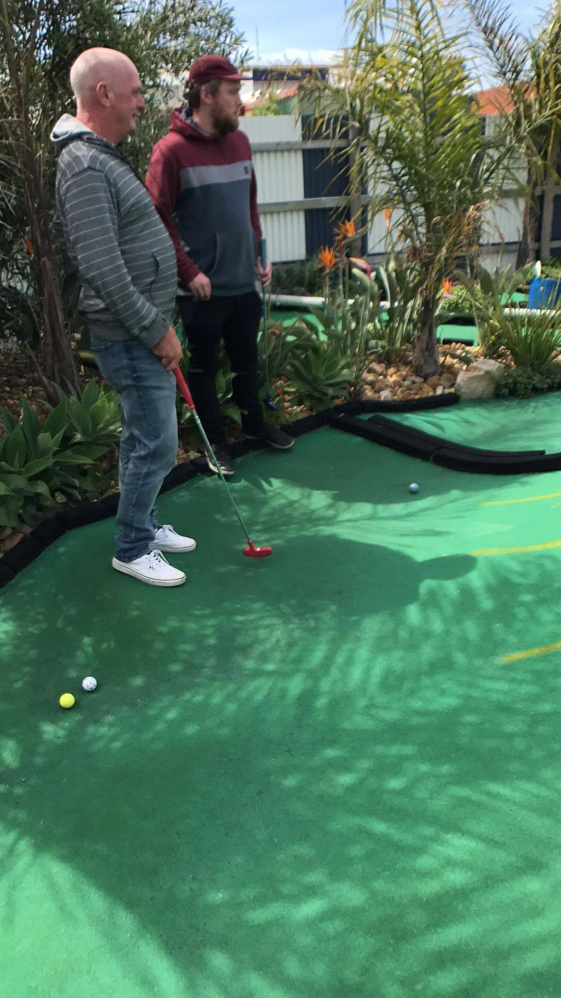 Two men playing mini golf outdoors on a green course, surrounded by plants and a garden fence, with one preparing to putt the ball.