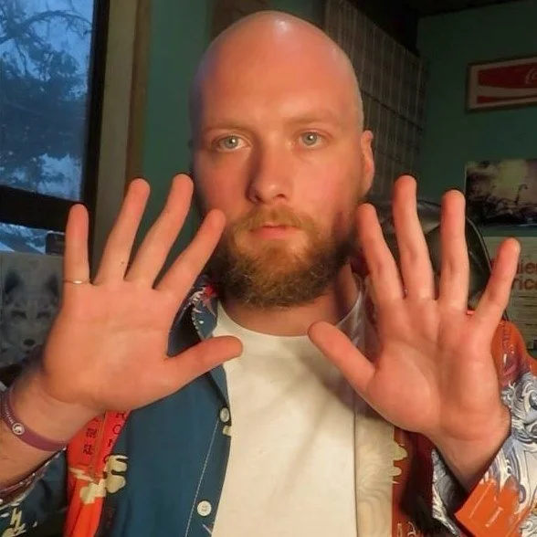 A man with a beard and bald head holding up both hands with open palms facing the camera indoors, with a window and posters on the wall in the background.