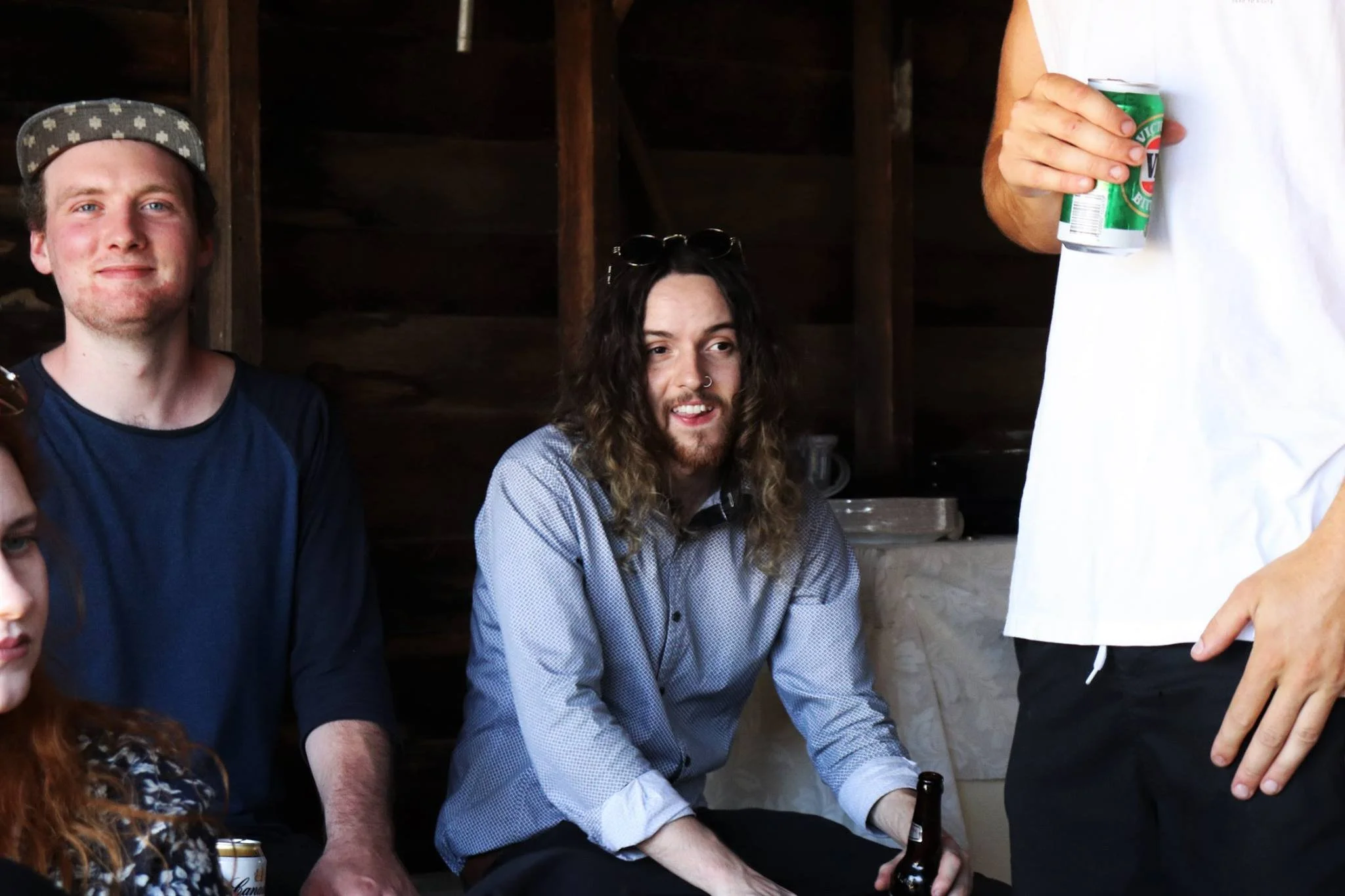 Group of young adults at a casual gathering, one holding a beer and another with sunglasses on their head, inside a wooden structure.