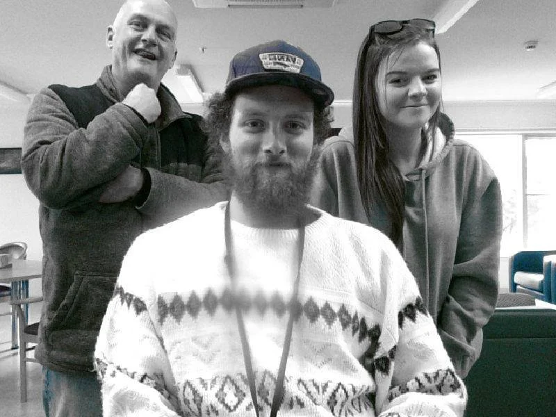 Three people smiling indoors. A man with a beard and a cap is sitting in front, a woman with long hair and glasses is on his right, and another man is standing behind them on the left, all casually dressed.