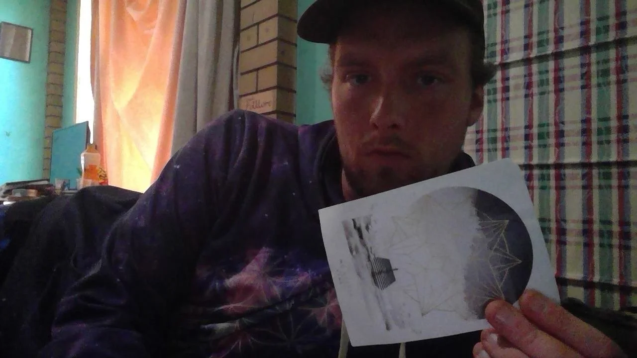 A young man with a cap holding a piece of paper featuring an image of a bridge with a spiderweb pattern in a room with curtains and a brick wall.