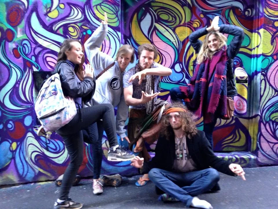 Five young people posing in front of a colorful, abstract mural. One person kneels on the ground, and others strike playful poses, with two standing and making various hand gestures, one sitting on a bench, all smiling or making quirky expressions.