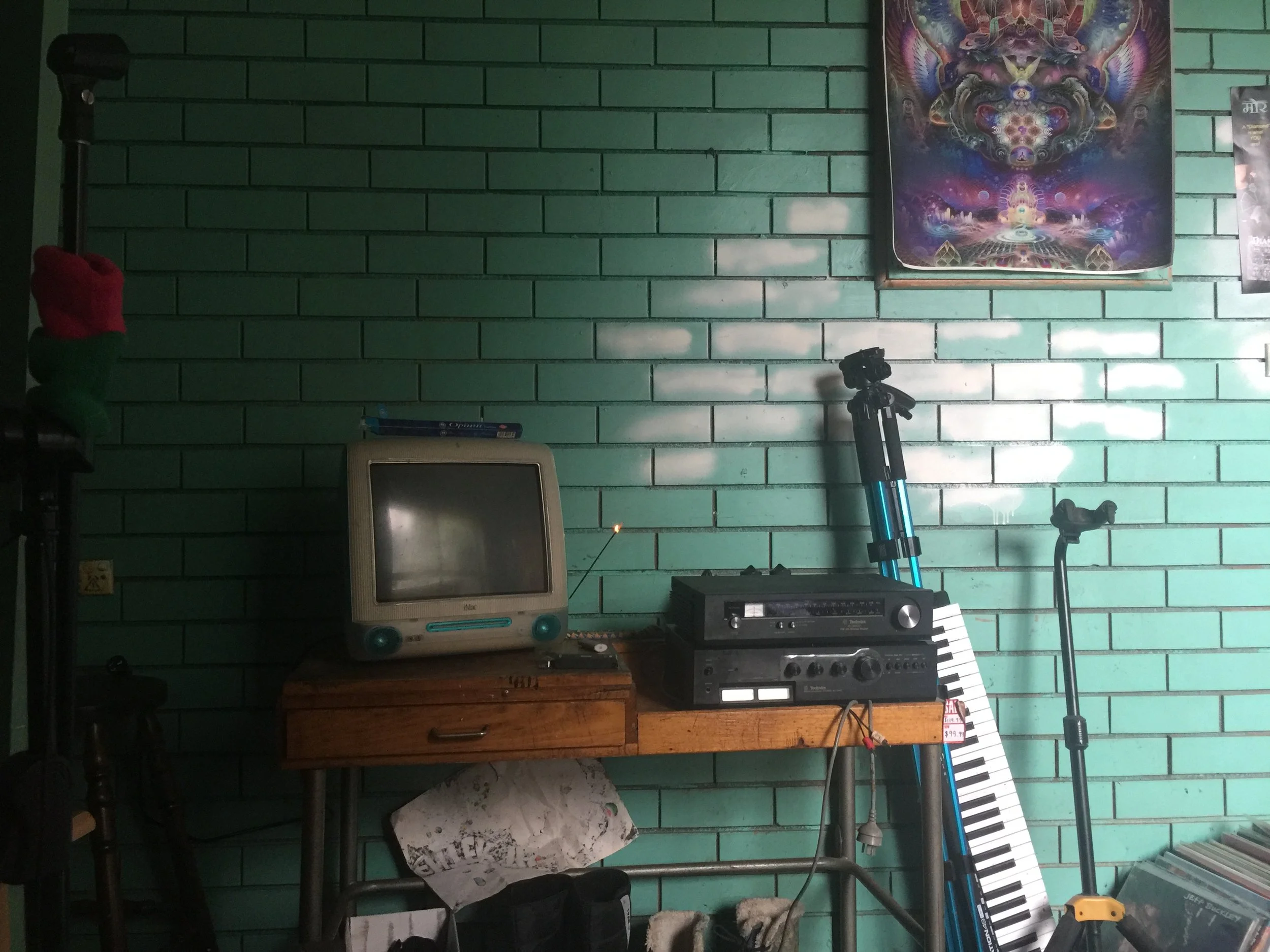 Room with green brick wall, vintage computer monitor on wooden table, electronic equipment, paint palette, and posters, with a keyboard, microphone, and camera on tripods.