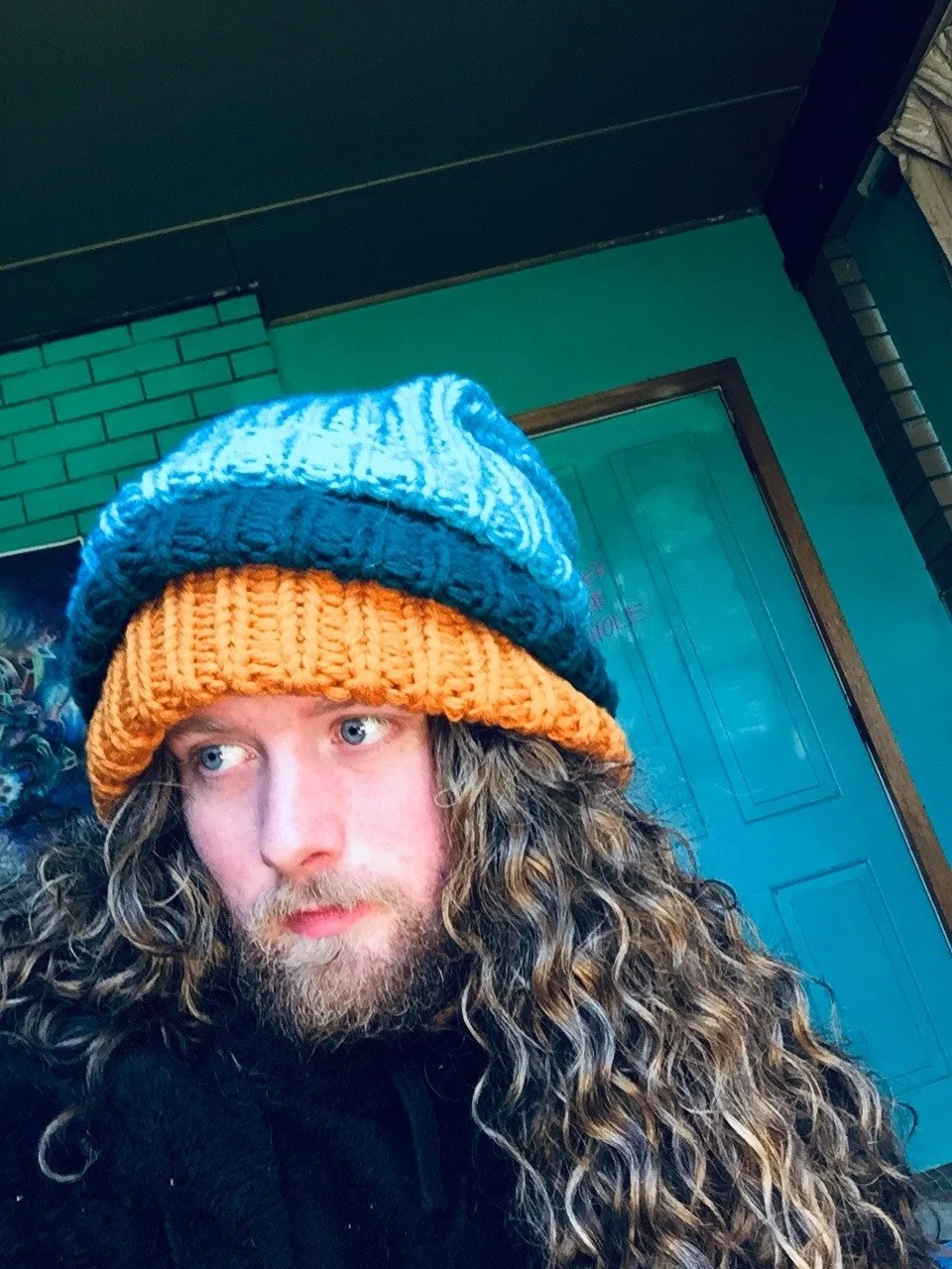 A man with long curly hair and a beard wearing a knit hat with blue and orange colors, indoors against a green and brick wall.