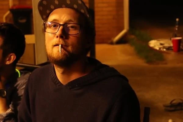 A man with glasses and a hat sitting outdoors at night, with a lit cigarette in his mouth.