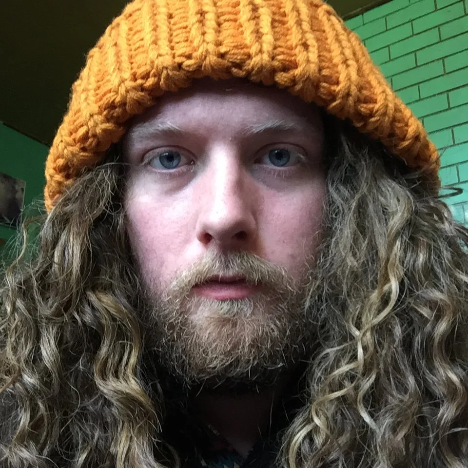 A person with long, curly hair, light skin, and a beard, wearing an orange knit beanie. The background includes a green brick wall.