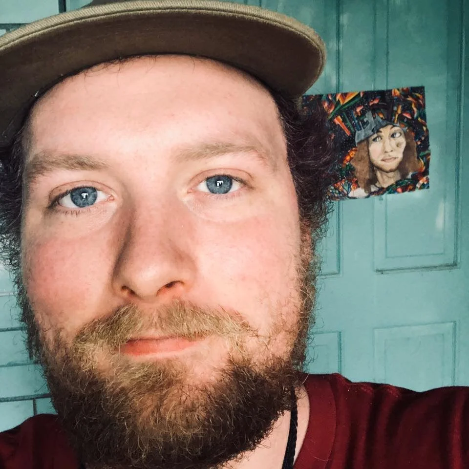 A man with blue eyes and a beard wearing a khaki baseball cap, taking a selfie in front of a green wall. Behind him, there is a colorful portrait of a woman with curly hair.